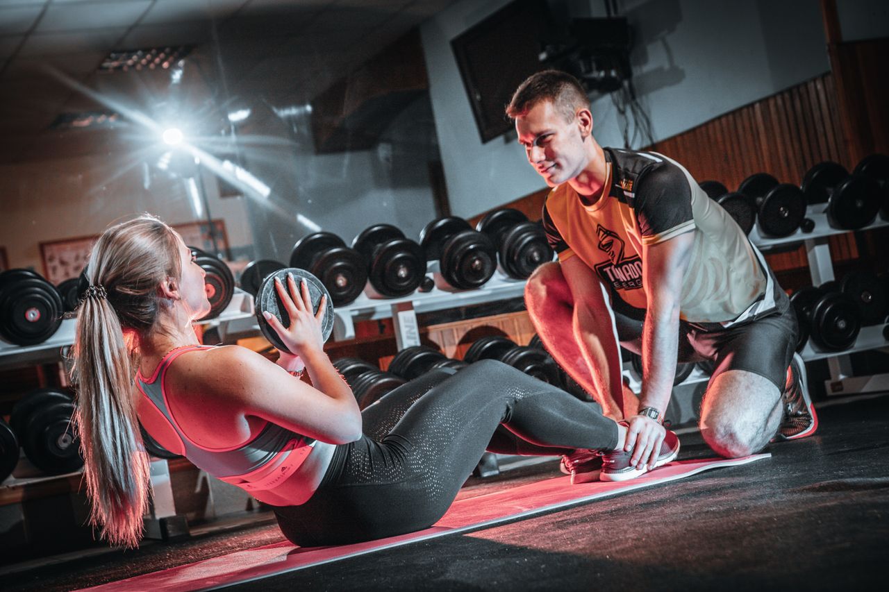 hero-img-01 A personal trainer assists a woman doing sit-ups with a medicine ball in a gym setting.